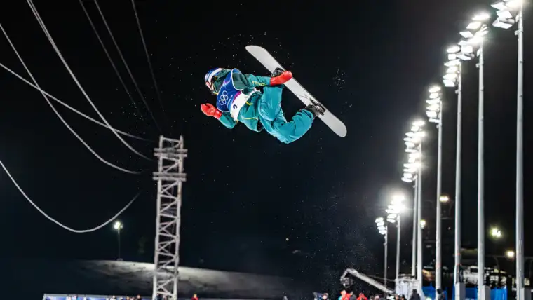 A male athlete wearing green Australian winter jacket and pants is mid-air performing a snowboard trick against a night sky.