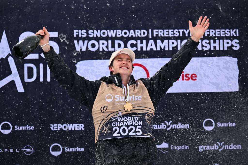A photo of a male in his thirties on top of a podium, wearing a black snow jacket, a 2025 World Champion bib, a gold medal around his neck and holding his arms up in triumph with a bottle of champagne in one hand.