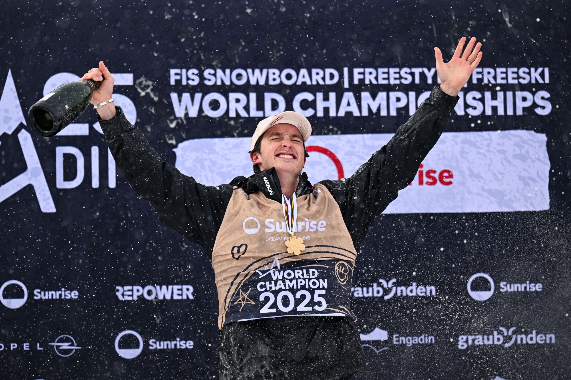 A photo of a male in his thirties on top of a podium, wearing a black snow jacket, a 2025 World Champion bib, a gold medal around his neck and holding his arms up in triumph with a bottle of champagne in one hand.