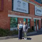 Two middle-aged women stand side-by-side in front of a red brick building. The lady on the left has dark hair and is wearing a light blue top, the lady on the right has light coloured hair with a white tee and navy blazer.