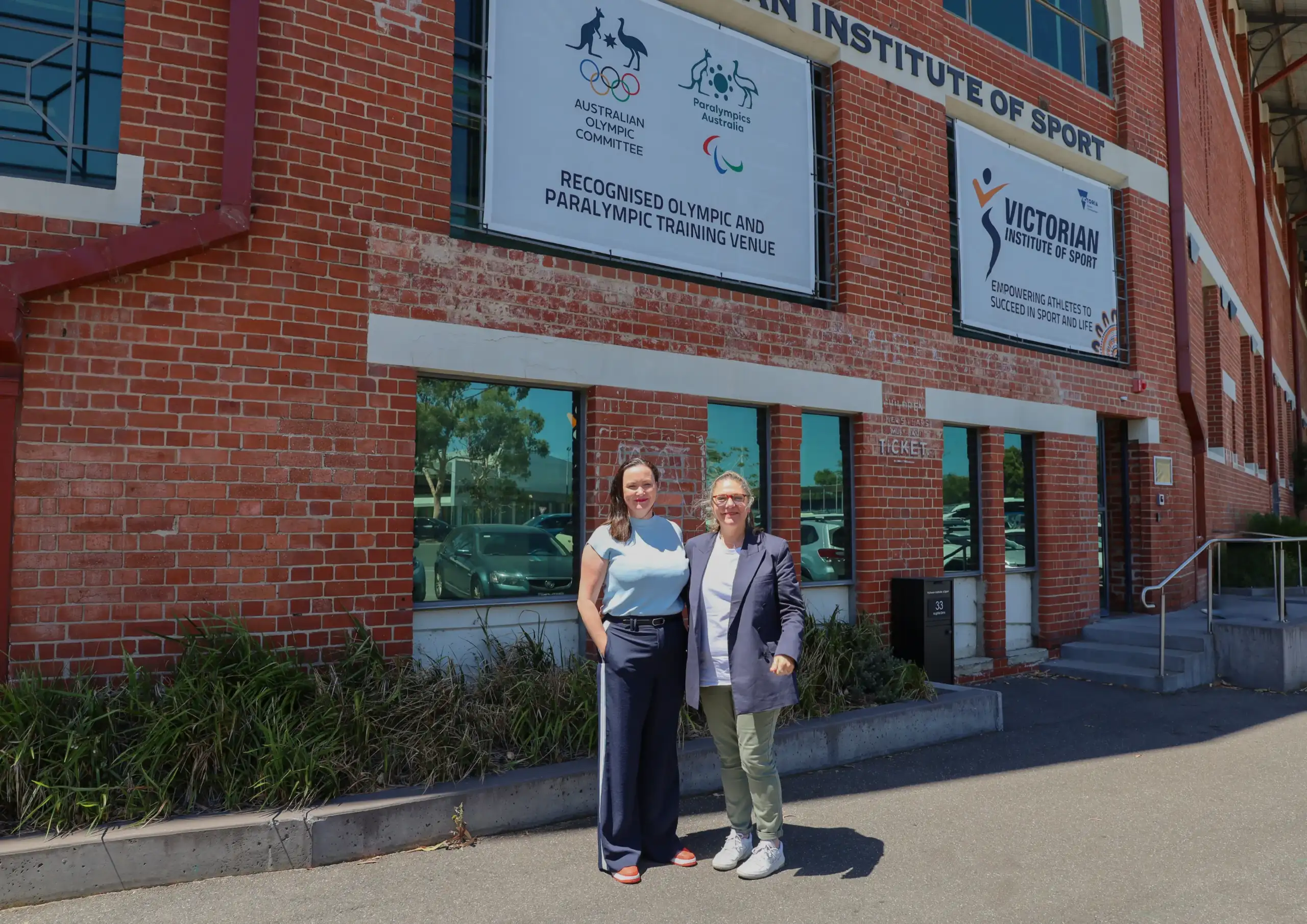VIS Board_Sarah Styles_Andrea Pearman 3 Two middle-aged women stand side-by-side in front of a red brick building. The lady on the left has dark hair and is wearing a light blue top, the lady on the right has light coloured hair with a white tee and navy blazer.