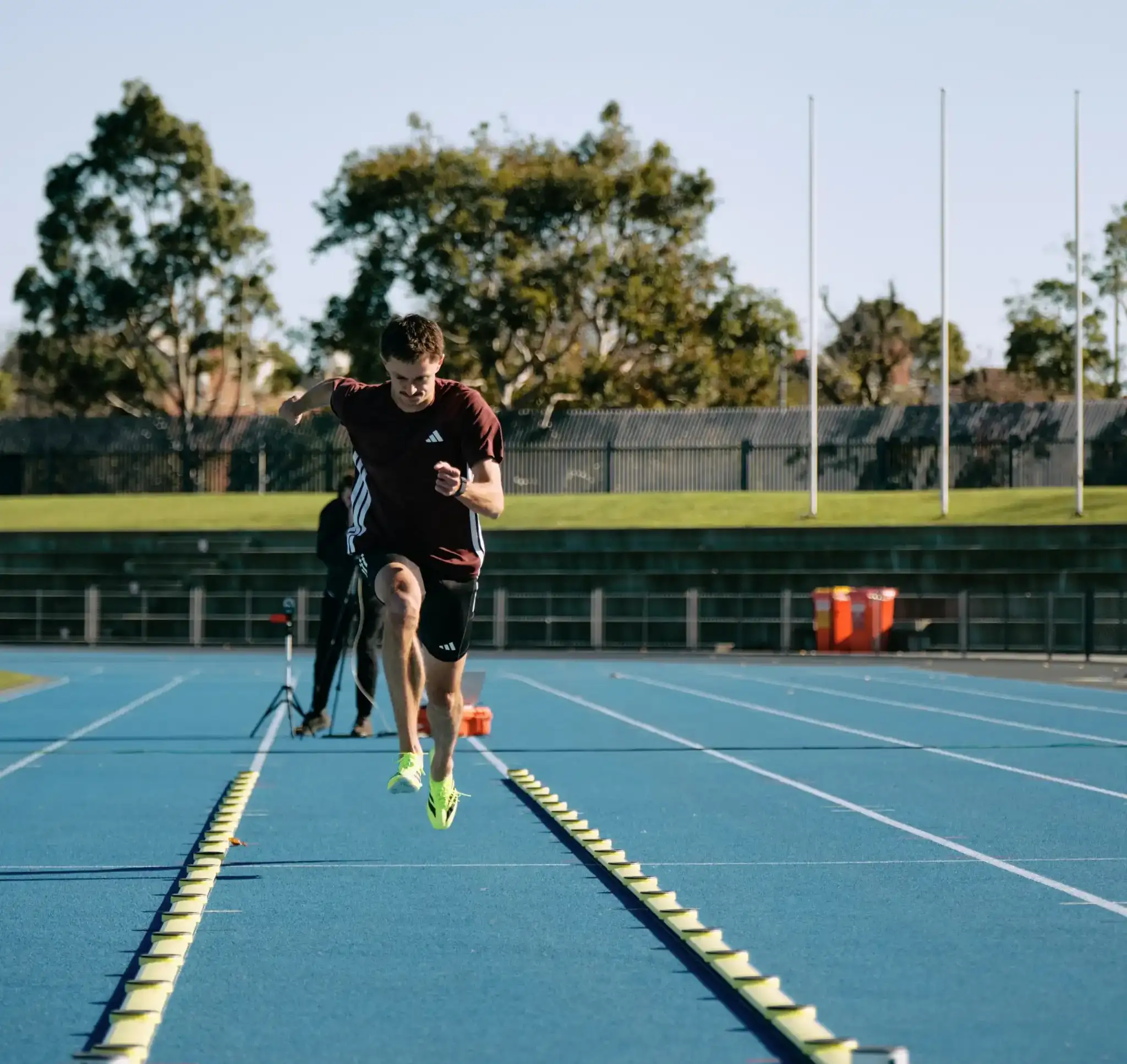 A male athlete is on an athletics track, sprinting towards the camera. Cones are set up on either side of his lane with a staff member and camera behind him filming. 