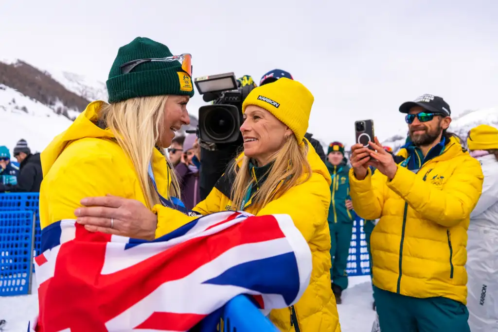 Two female athletes hold each others arms in celebration, both wearing yellow Australian Olympic Team snow jackets.