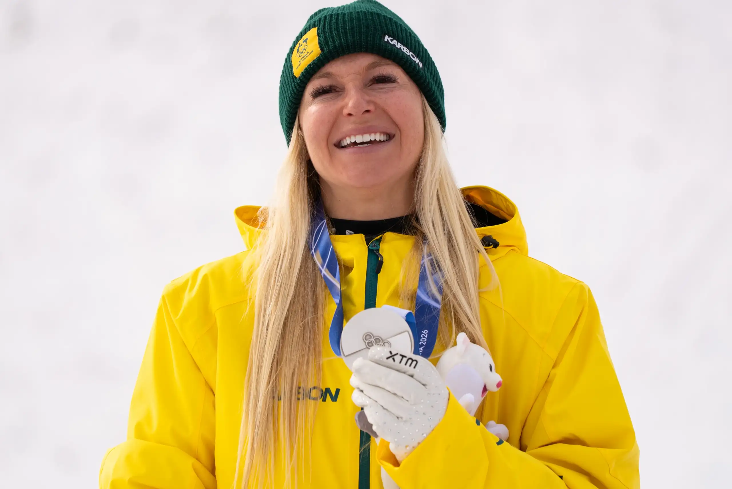 A female athlete with long straight blonde hair is wearing a green beanie and a yellow Australian Olympic Team snow jacket. She has a silver medal around her neck.