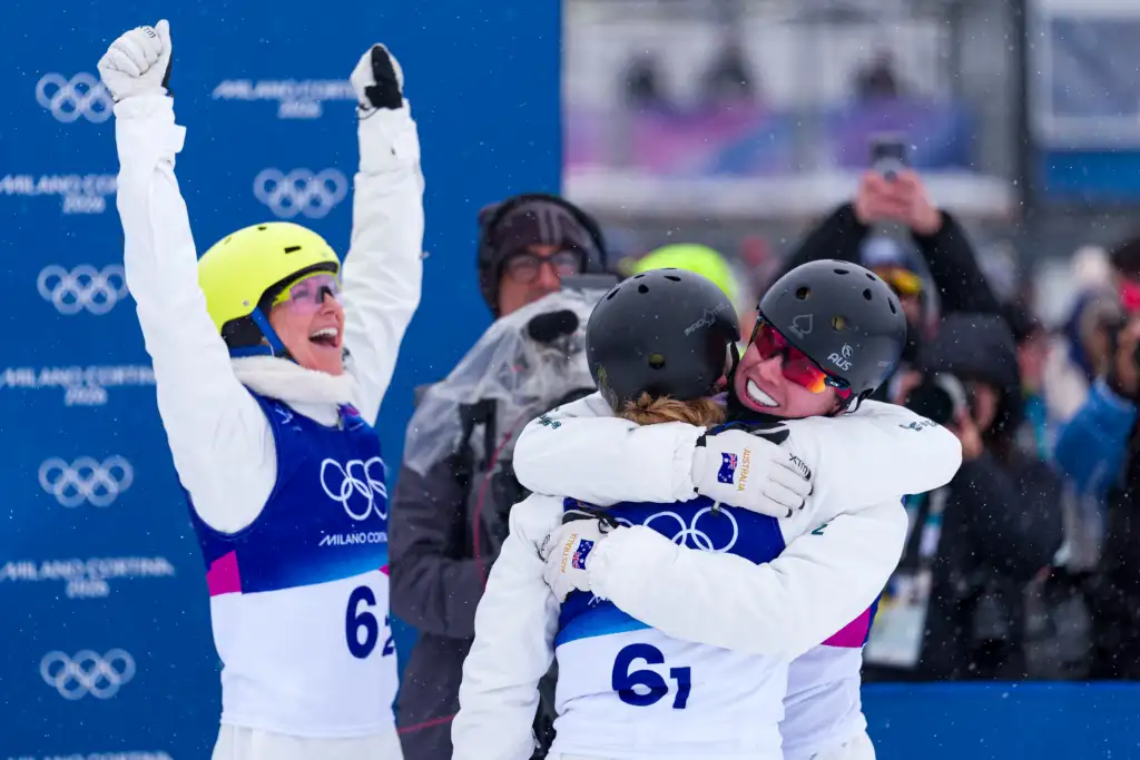 A photo of three athletes wearing white snow jumpsuits and celebrating. A female on the left wears a bright yellow helmet and has her arms up in triumph. A female and male on the right, both wearing black helmets are hugging. All athletes wear a Milano Cortina competition bib which is blue.