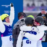 A photo of three athletes wearing white snow jumpsuits and celebrating. A female on the left wears a bright yellow helmet and has her arms up in triumph. A female and male on the right, both wearing black helmets are hugging. All athletes wear a Milano Cortina competition bib which is blue.