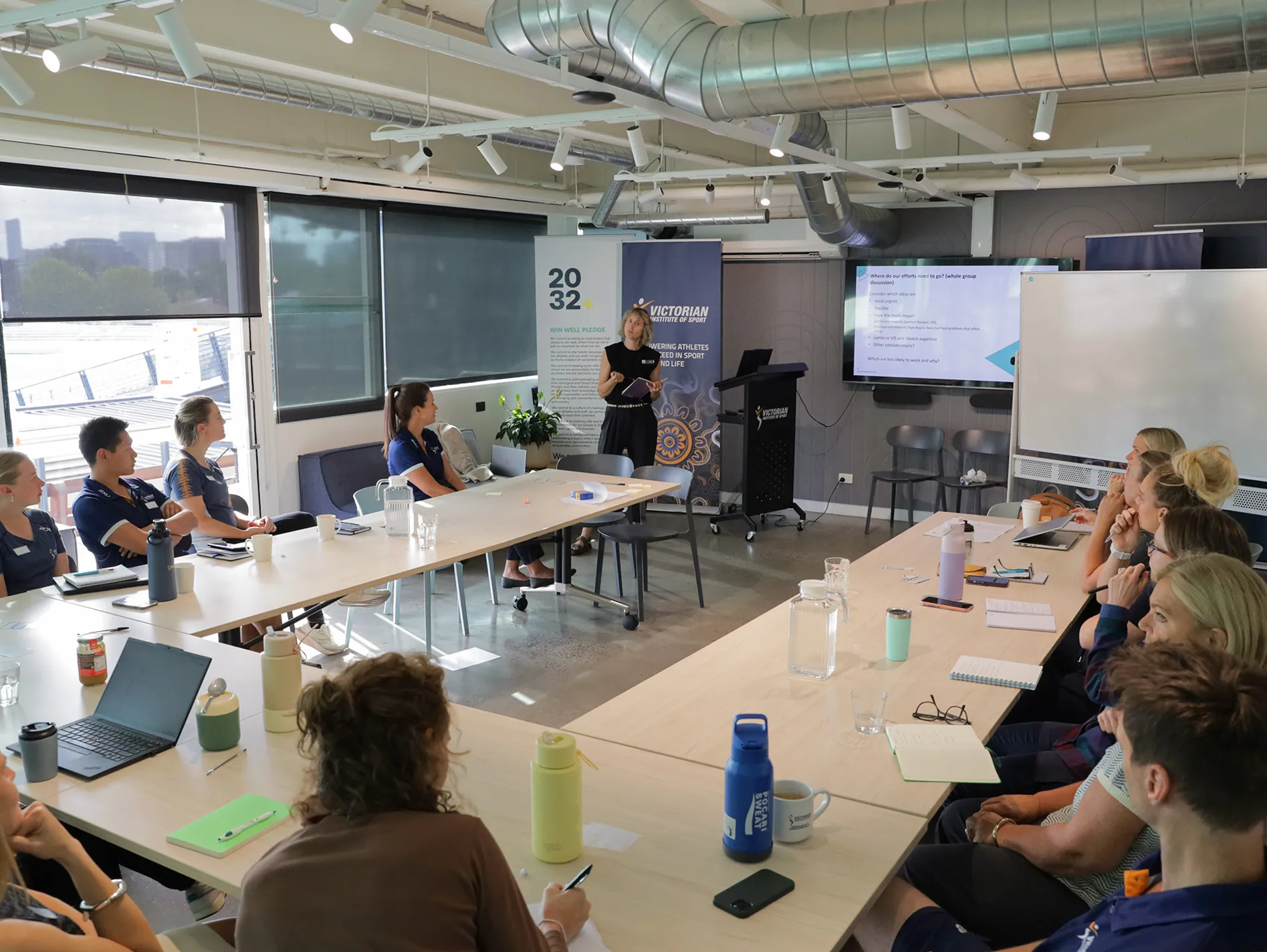A female in her twenties with brown hair and in a black top, is standing on stage behind a lectern presenting. 