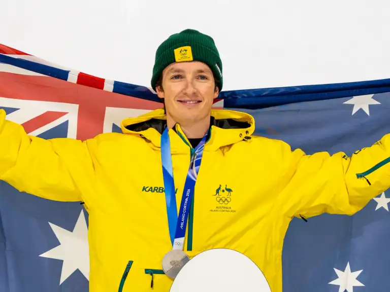 A male athlete in his thirties wearing a yellow Australian Olympic Team snow jacket, is wearing a silver Olympic medal around his neck and holding up the Australian flag behind him.