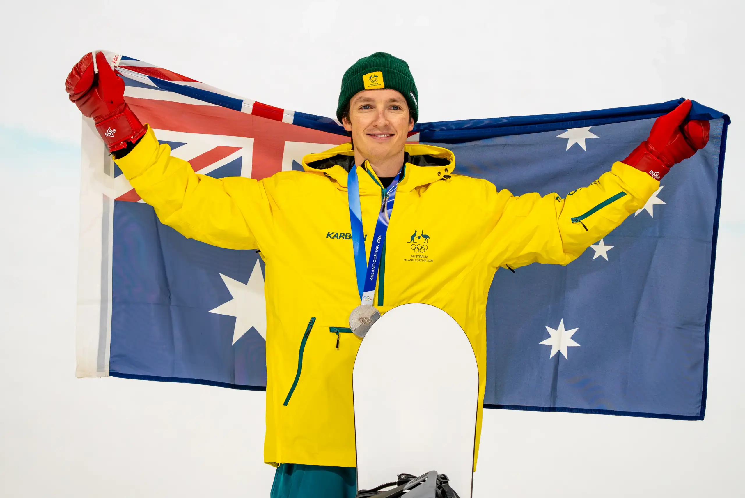 A male athlete stands on a podium wearing a yellow Australian Winter Olympic Team snow jacket. He has a silver medal around his neck and is holding up the Australian flag behind him.