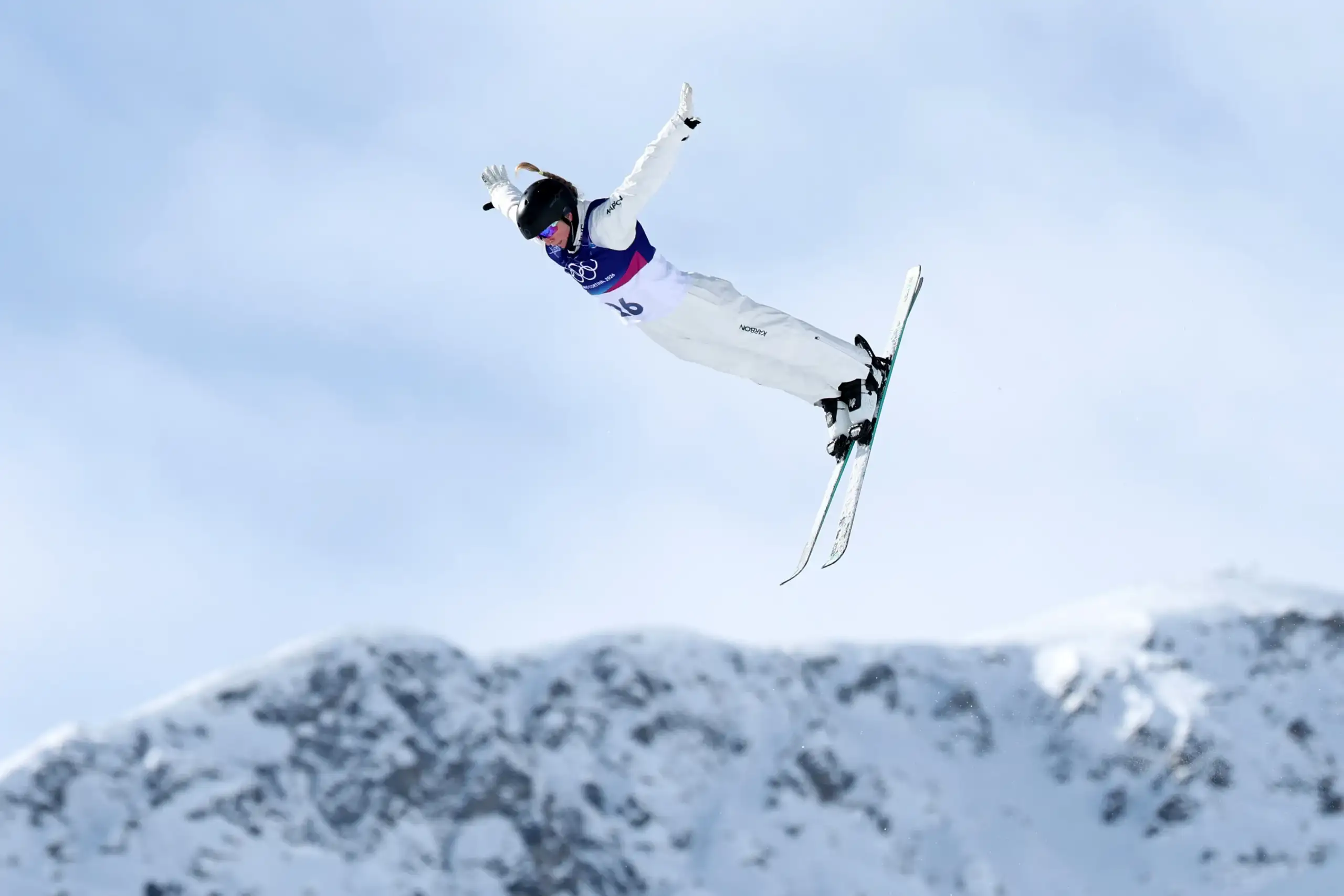 VIS - Sidney Stephens competing at Milano Cortina 2026 A woman wearing a white snow jumpsuit and skis flies through the air with a blue sky and snowcapped mountains in the background.