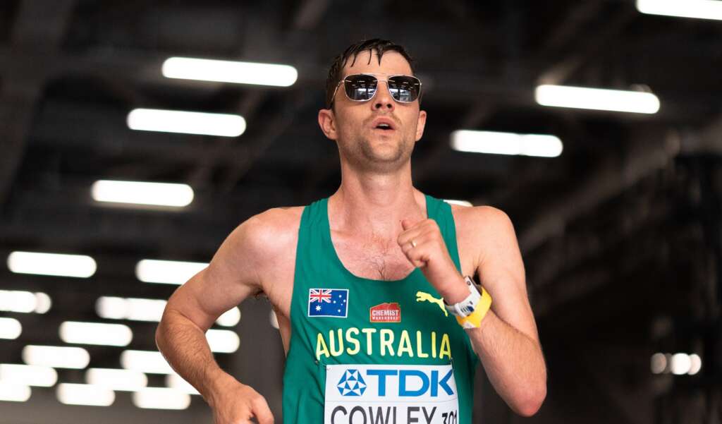 A male athlete with short dark hair wears a green Australian Team singlet, sunglasses and is in action mid race walk.