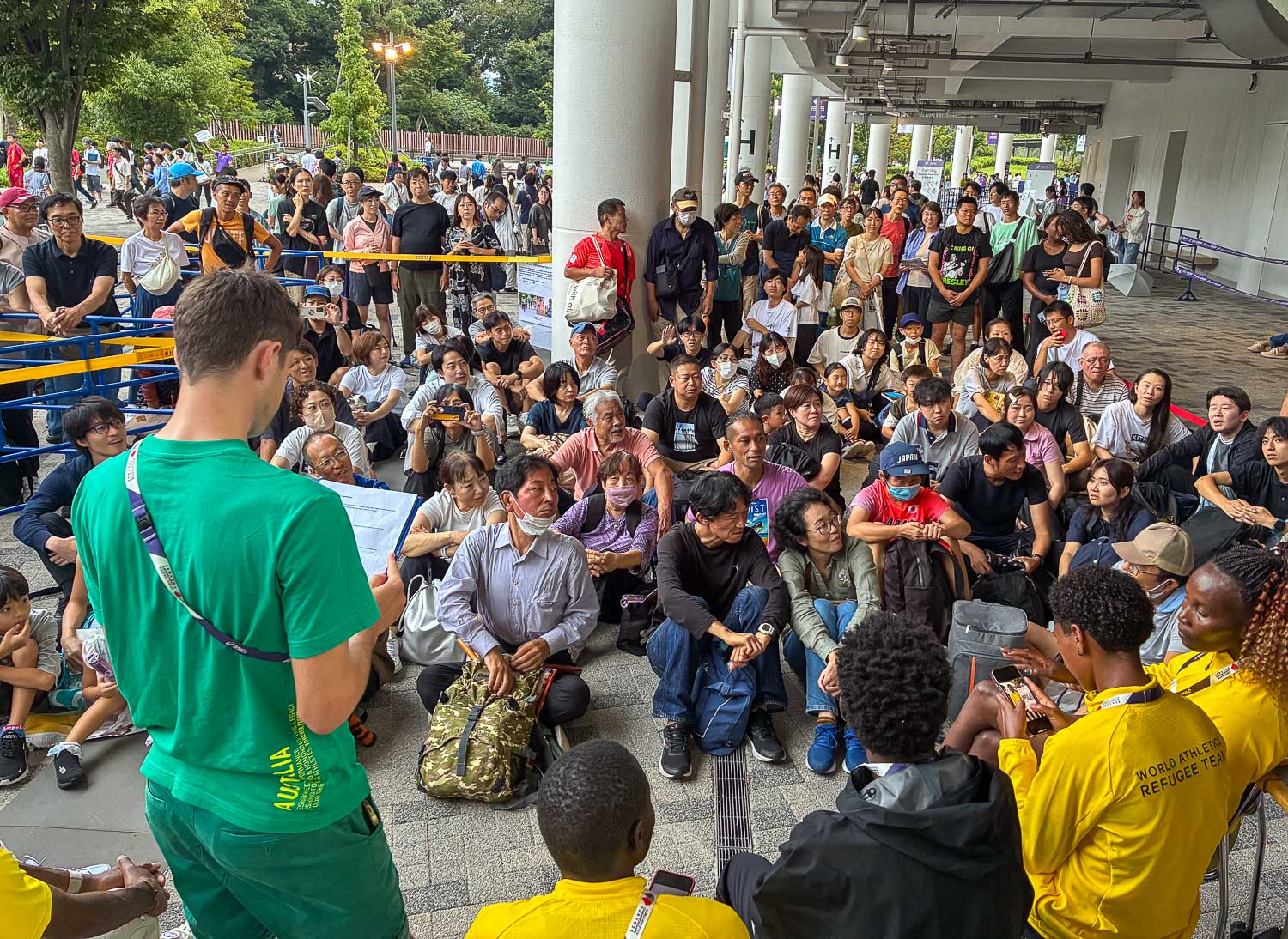 A wide angle photo of a large group of a Japanese school group, listening to a male athlete with short brown hair wearing green Australian Team tee and shorts, presenting to the group.