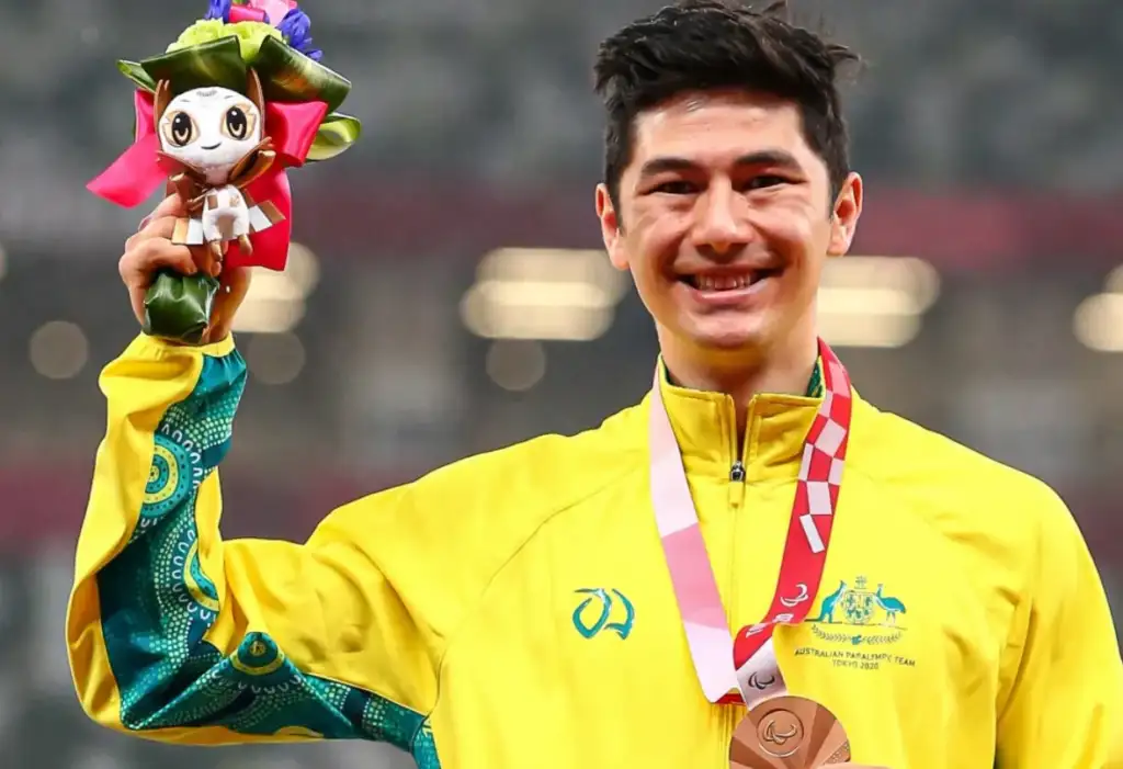 A male athlete in his thirties wearing a yellow Australian Paralypmic Team uniform, holding a bronze medal and a toy in his hand.