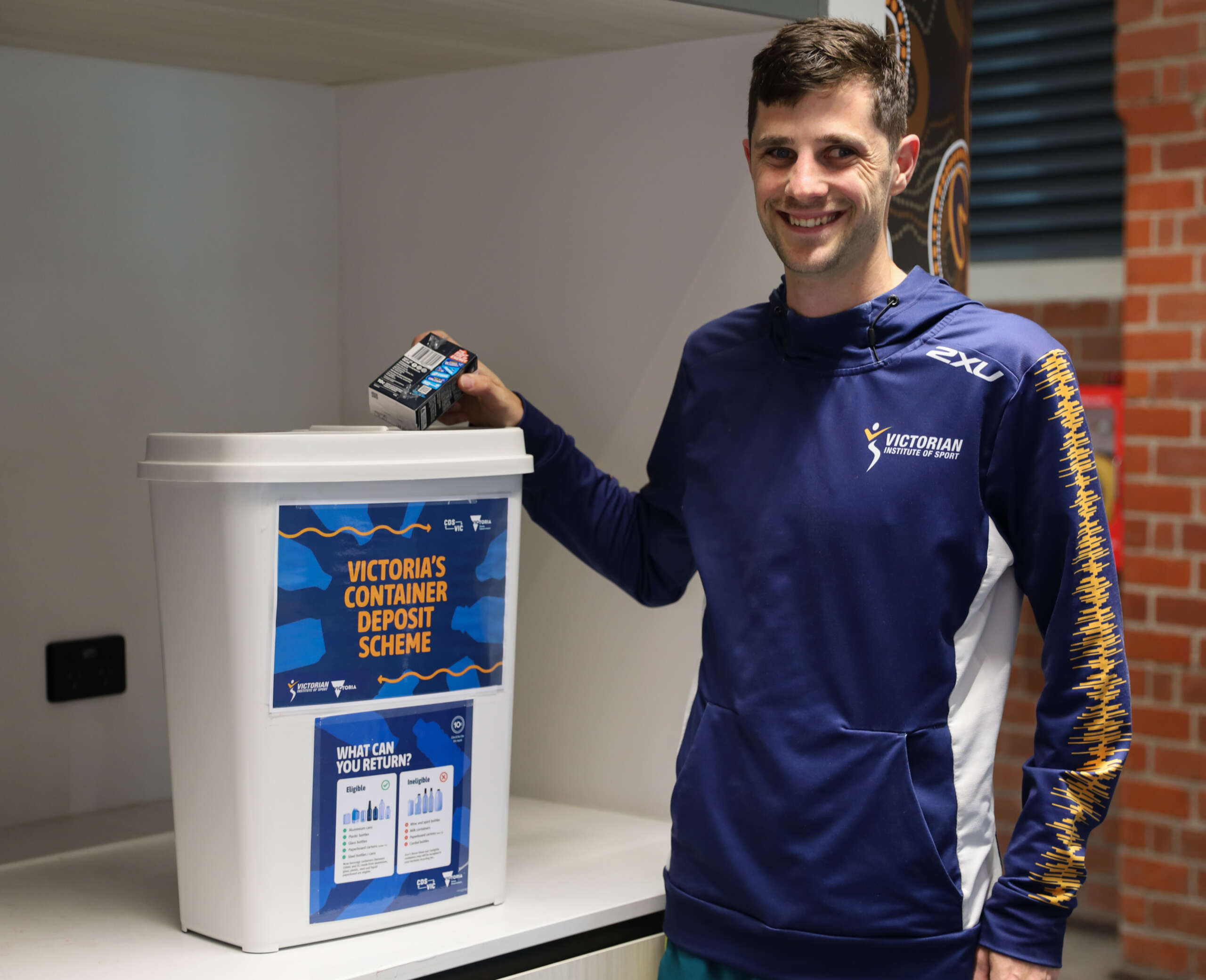 A male athlete with short dark hair wearing a VIS jumper is putting recycling into a bin with a sign reading Victorian Container Deposit Scheme.