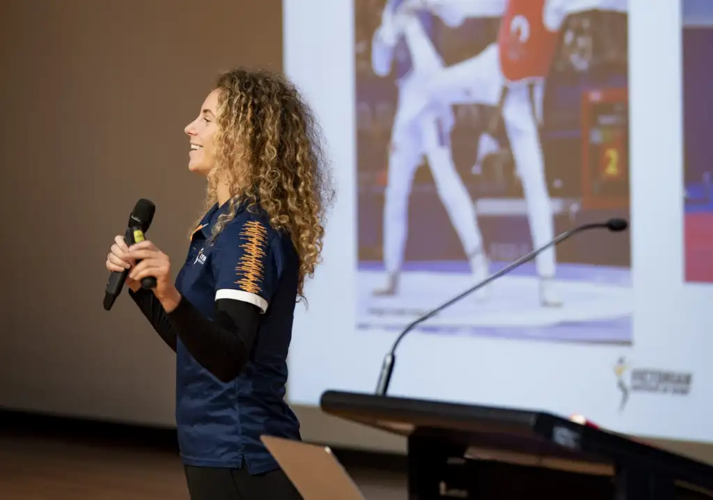 A female athlete in her twenties with long curly hair is presenting holding a microphone with a presentation in the background.
