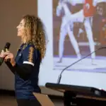 A female athlete in her twenties with long curly hair is presenting holding a microphone with a presentation in the background.
