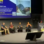 Four people sit on a large stage in a panel conversation with an AIS presentation in the background.