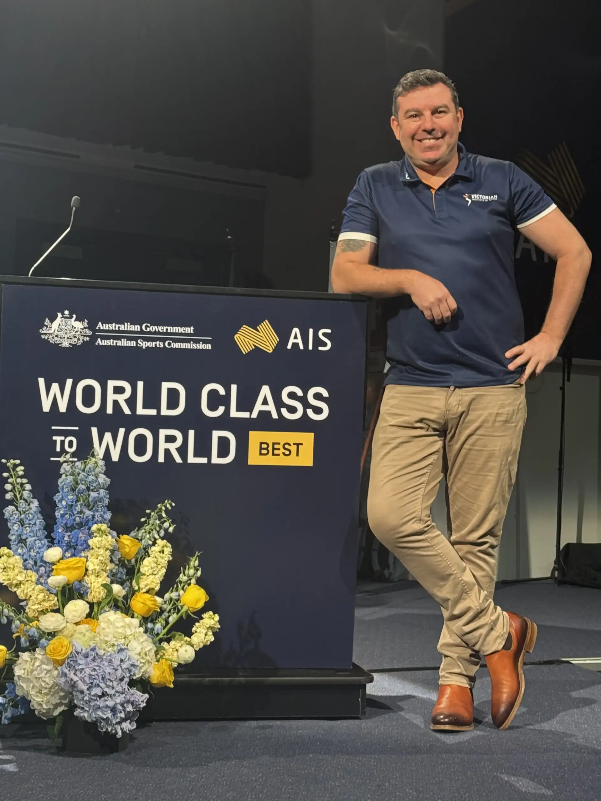 A man in his fourties with short dark hair and wearing a VIS polo, stands on a stage leaning against a large lectern reading 'World Class to World Best'.