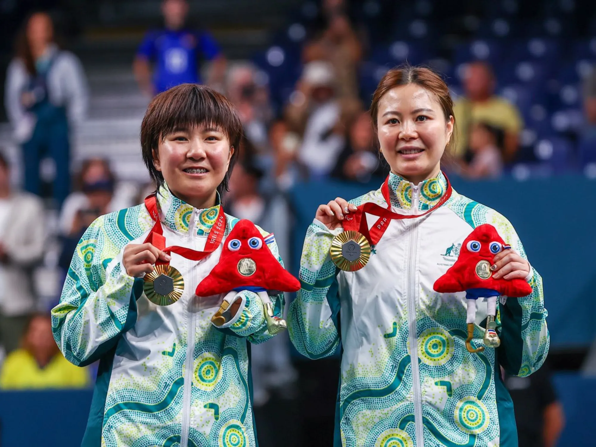 Two female Para athletes wearing Australian Paralympic uniform stand on a podium holding up gold medals and Paris mascot toys.
