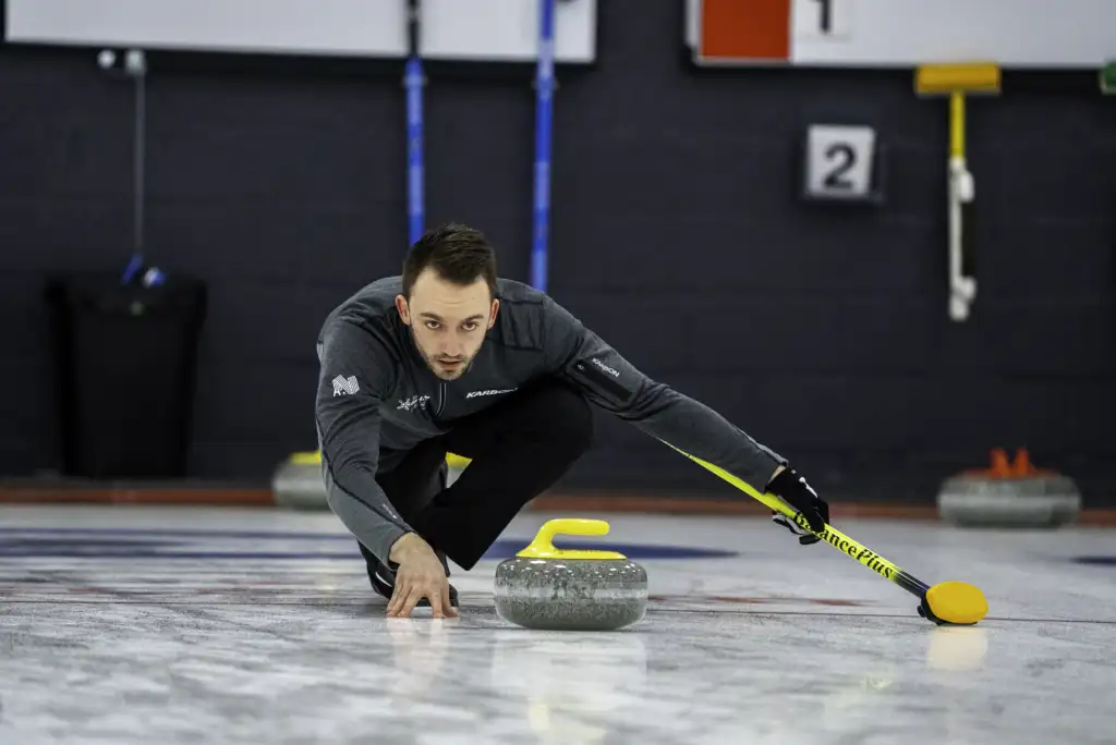 A photo of a male athlete with short dark hair in a lunge position, on ice, looking at a grey curling stone.