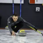 A photo of a male athlete with short dark hair in a lunge position, on ice, looking at a grey curling stone.