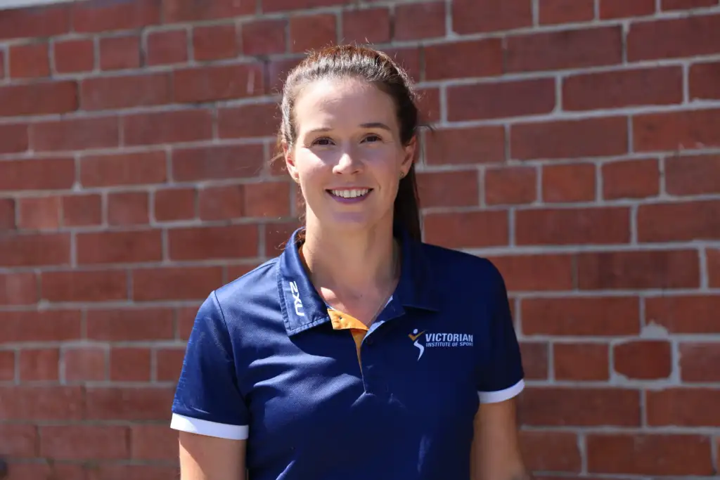 A photo of a Caucasian female smiling to the camera, she has brown hair tied up into a ponytail and is wearing a navy blue VIS polo. She stands in front of a brick wall.