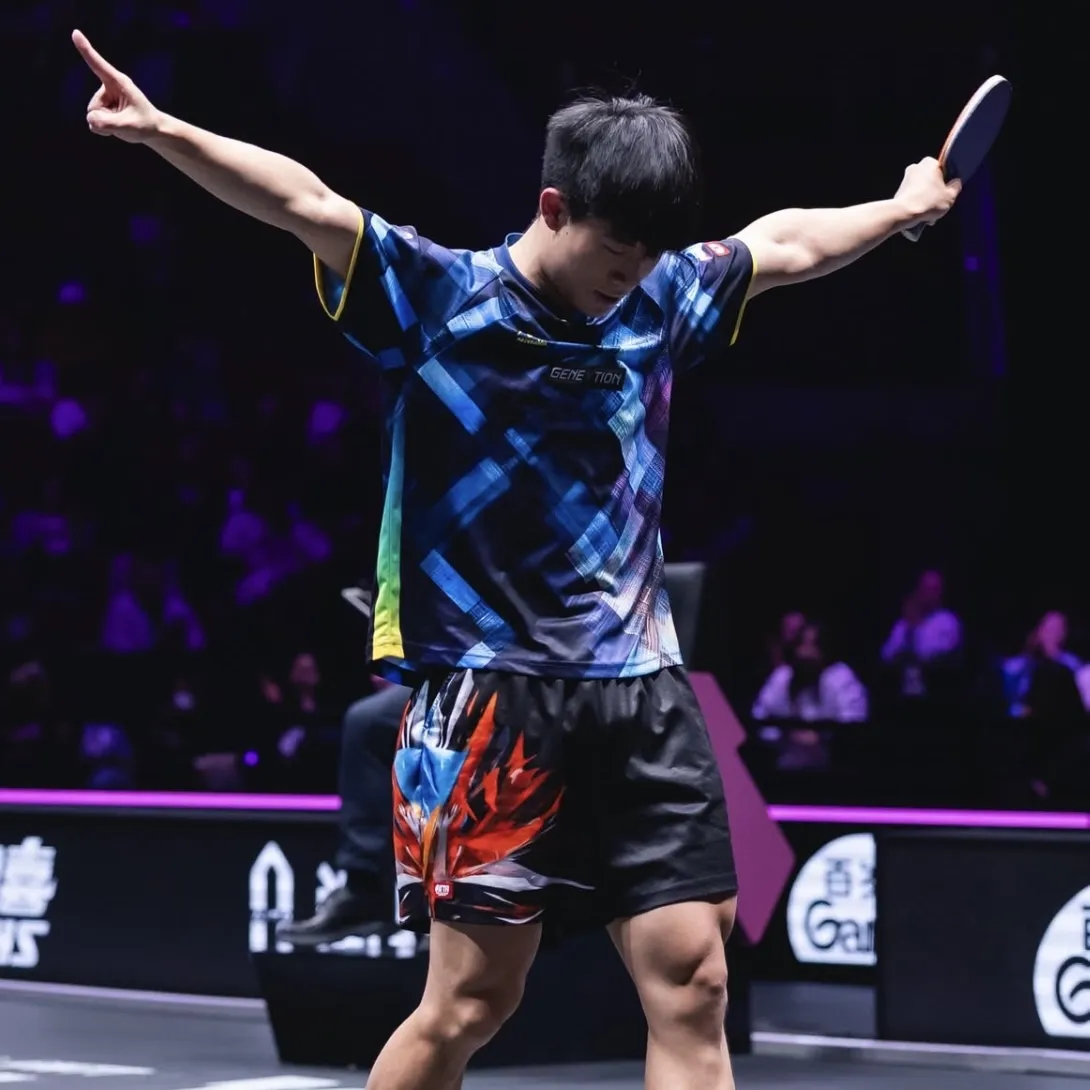 A young male athlete with short dark hair is walking with his arms out wide in triumph. He has a table tennis bat in his left hand and a competition crowd is blurred in the background.