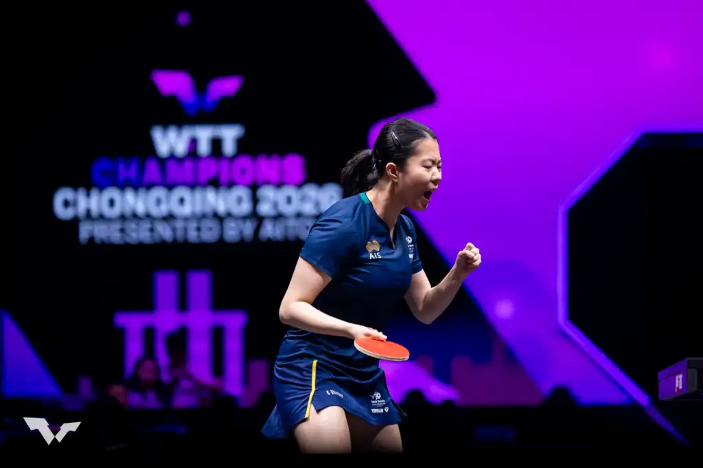 A female athlete pumps her fist in the air in triumph. She holds a table tennis bat in her right hand and purple and black competition branding is in the background.