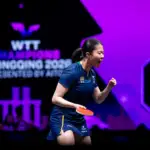 A female athlete pumps her fist in the air in triumph. She holds a table tennis bat in her right hand and purple and black competition branding is in the background.