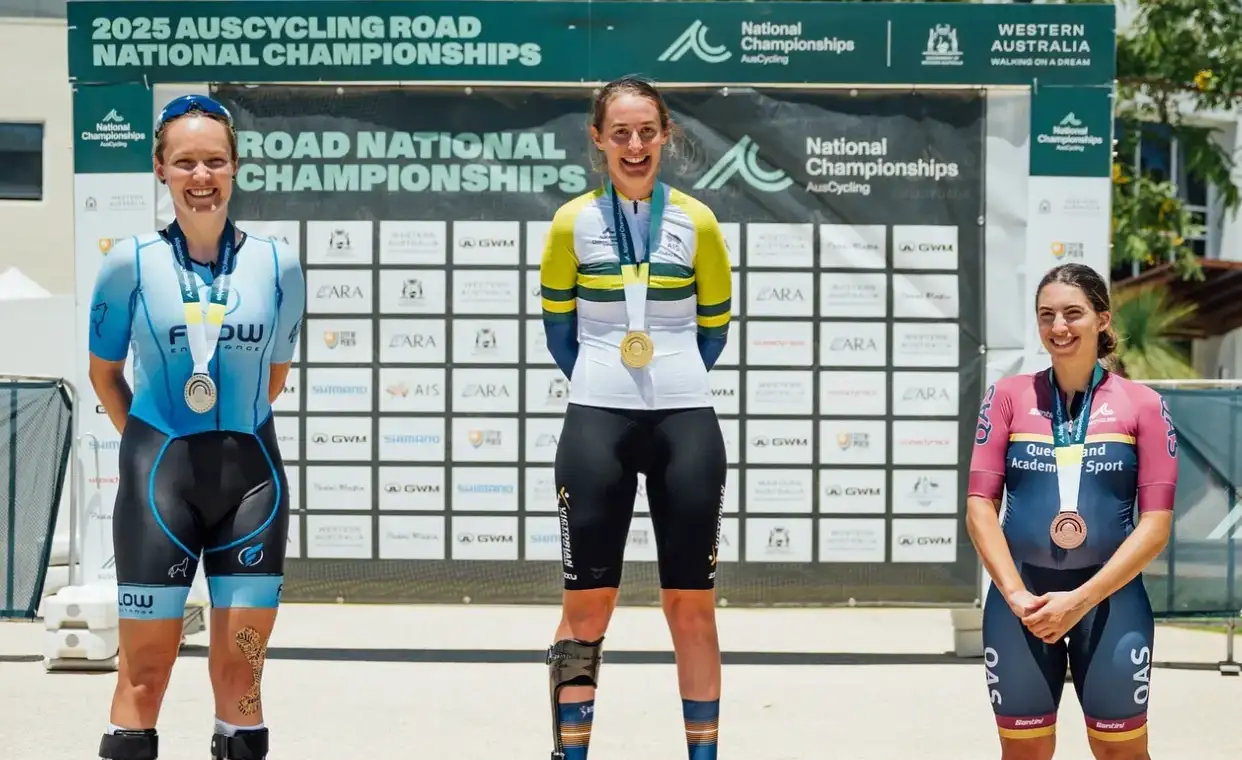Three female athletes stand on a podium wearing cycling lycra. Behind them is a media banner reading Road National Championships. The middle female athlete standing atop the podium wears a white, green and gold jersey indicating National Champion. 