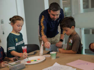 A man demonstrates to two children how to paint their boomerangs.
