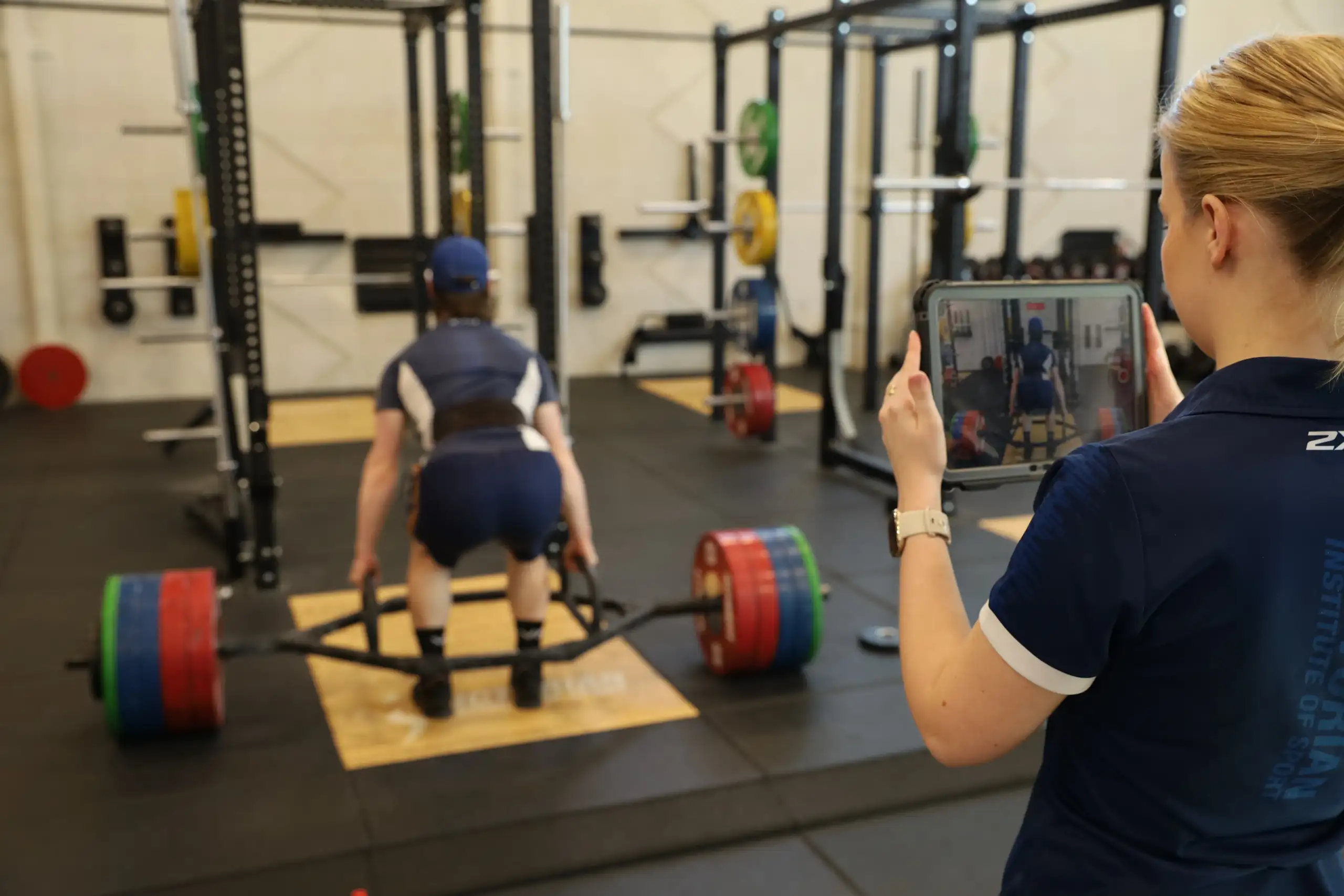 A strength training session in a gym. One person performs a deadlift with a barbell loaded with coloured weight plates while standing on a lifting platform inside a power rack. In the foreground, another person records the lift on a tablet. Additional strength training equipment is visible throughout the gym.