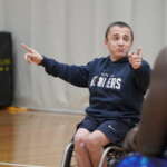 A man with short hair wearing a navy sweatshirt and shorts in a wheelchair. Pointing to other wheelchair basketball players as he coaches.