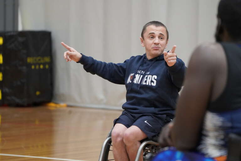 A man with short hair wearing a navy sweatshirt and shorts in a wheelchair. Pointing to other wheelchair basketball players as he coaches.