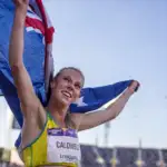 A female athlete with tied up blonde hair wears a yellow running crop top with a competition bib pinned to the front with CALDWELL written on the front. She holds up an Australian flag behind her and smiles into the distance.