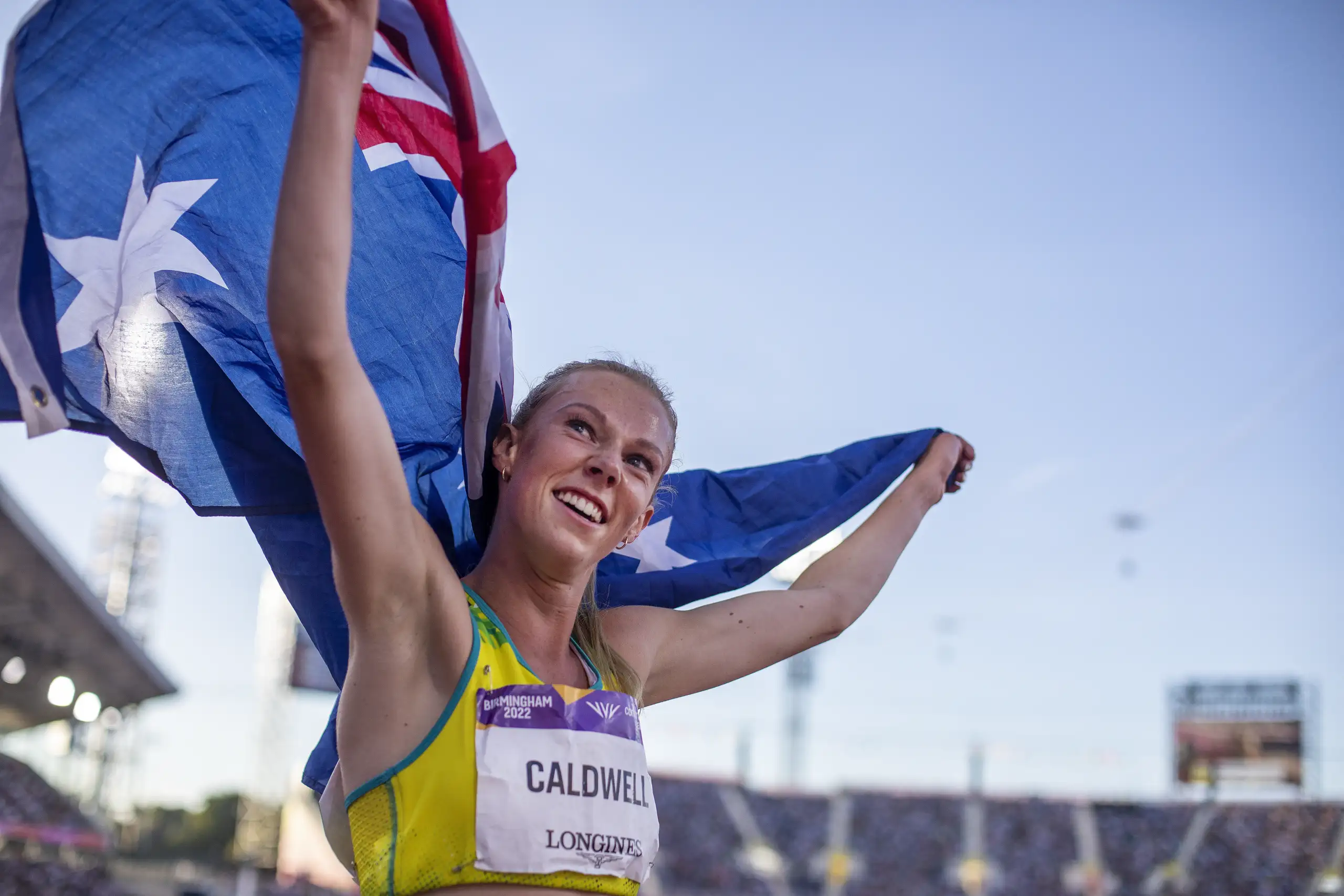 A female athlete with tied up blonde hair wears a yellow running crop top with a competition bib pinned to the front with CALDWELL written on the front. She holds up an Australian flag behind her and smiles into the distance.