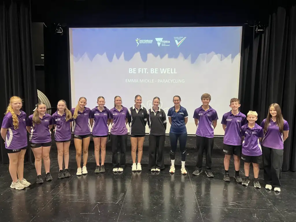 A group of high school students all wearing purple tops and black shorts stand in a line smiling to camera. In the middle is a female athlete wearing a navy VIS polo, with a grey and navy projection screen in the background reading Be Fit. Be Well.