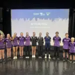 A group of high school students all wearing purple tops and black shorts stand in a line smiling to camera. In the middle is a female athlete wearing a navy VIS polo, with a grey and navy projection screen in the background reading Be Fit. Be Well.