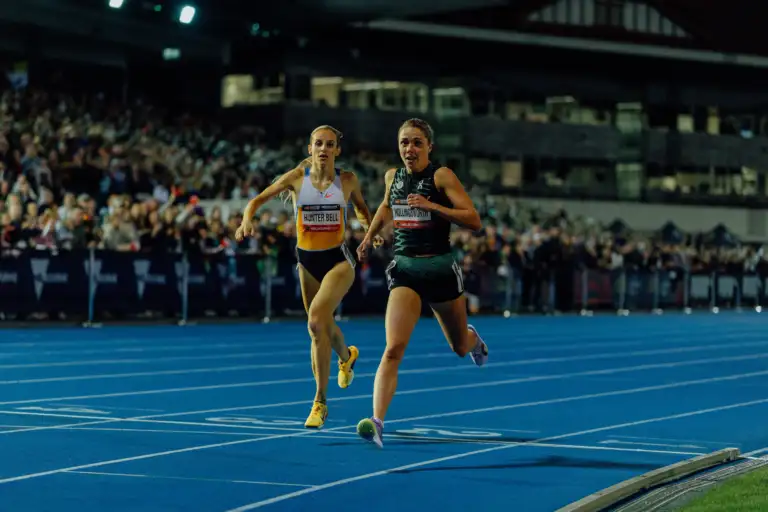 Two female athletes are running across the finish line on a blue athletics track. A large crowd is in the background and they're in action for a close finish.