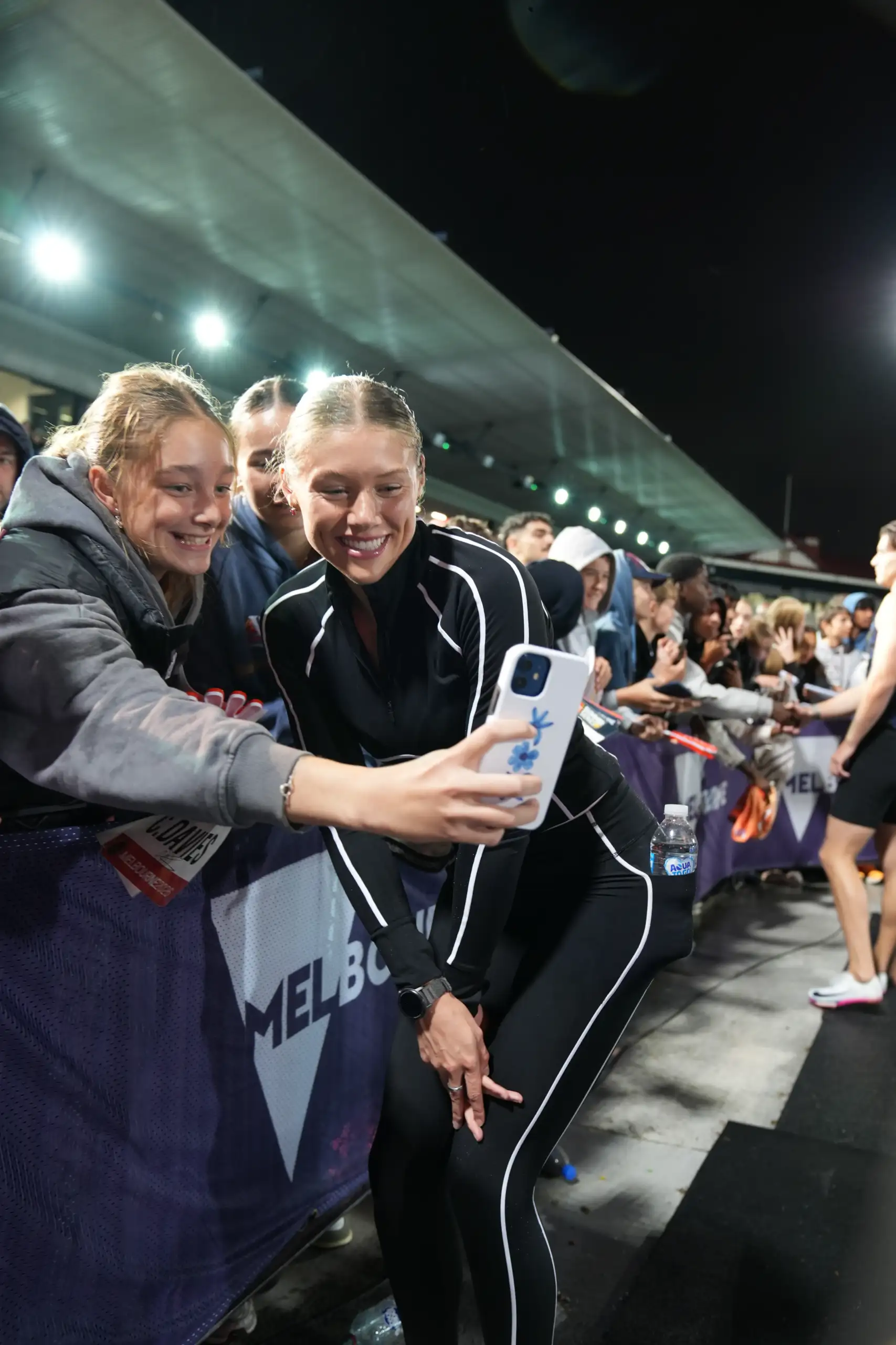 A female athlete with blonde hair tied back, wears a long sleeved black top and long leggings. She is smiling for a photo with a young girl in the crowd who is holding out an iPhone for a selfie.