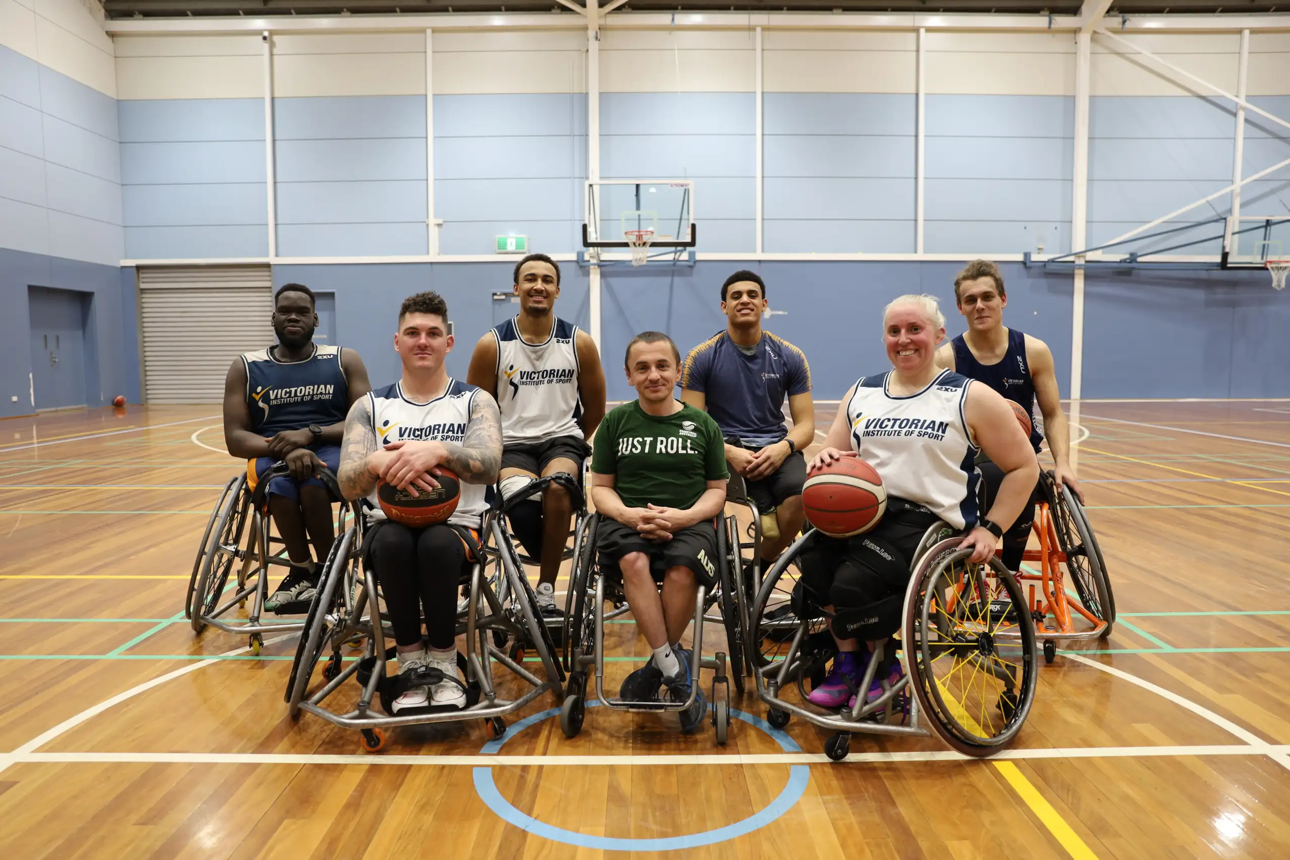 A group of 6 male and 1 female wheelchair basketball players posing for a team picture on an indoor basketball court.