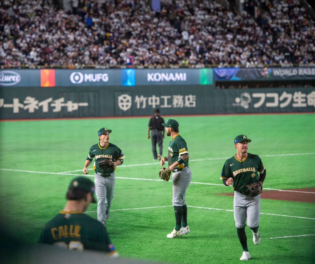 Three male baseball players stand talking on green field in Australia uniform wearing baseball gloves. 