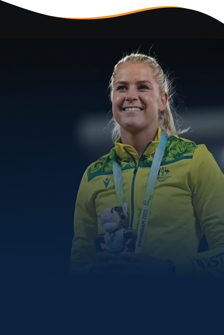 A photo of a female athlete with blonde hair tied into a ponytail. She wears a yellow zip up fleece from the Australian Commonwealth Games Team. She smiles into the distance with a medal around her neck and holding a mascot toy in front of her chest.