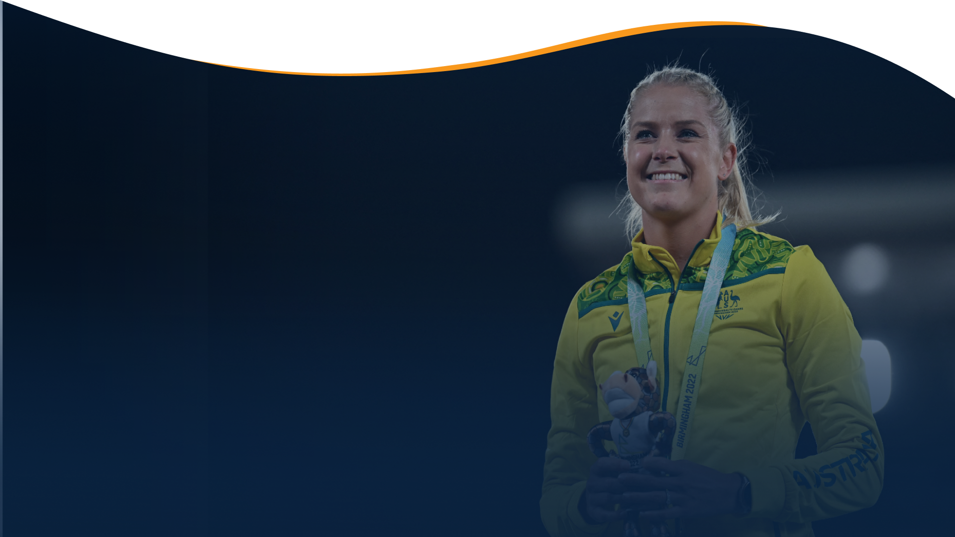 A photo of a female athlete with blonde hair tied into a ponytail. She wears a yellow zip up fleece from the Australian Commonwealth Games Team. She smiles into the distance with a medal around her neck and holding a mascot toy in front of her chest.