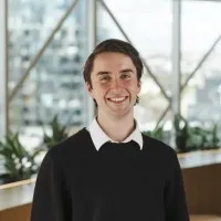 A professional headshot of a young Caucasian male with short dark brown hair. He wears a black jumper with a white collar tucked out. Behind him are some indoor plants and large windows with city buildings in the background.