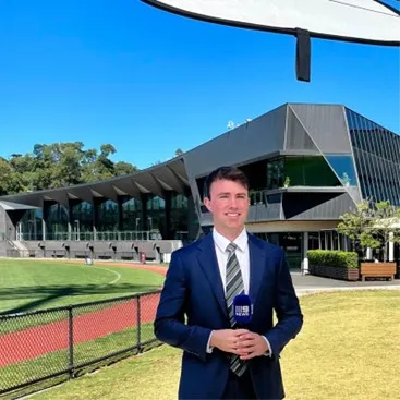 A young Caucasian male with short brown hair, he stands holding a Channel 9 microphone in front of a large black corporate building next to a football oval.