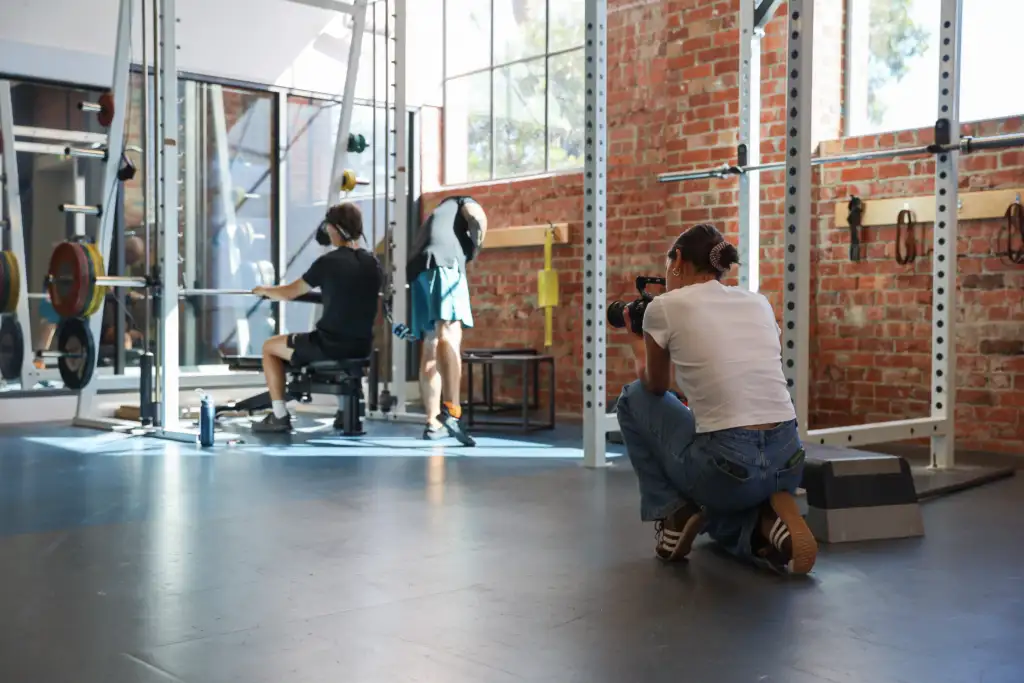 The photo is taken in a gym, in the background is an athlete performing an exercise. In the foreground is the back of a young female in a white t-shirt and blue jeans, kneeling with a camera in her hands facing the athlete.