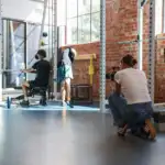 The photo is taken in a gym, in the background is an athlete performing an exercise. In the foreground is the back of a young female in a white t-shirt and blue jeans, kneeling with a camera in her hands facing the athlete.