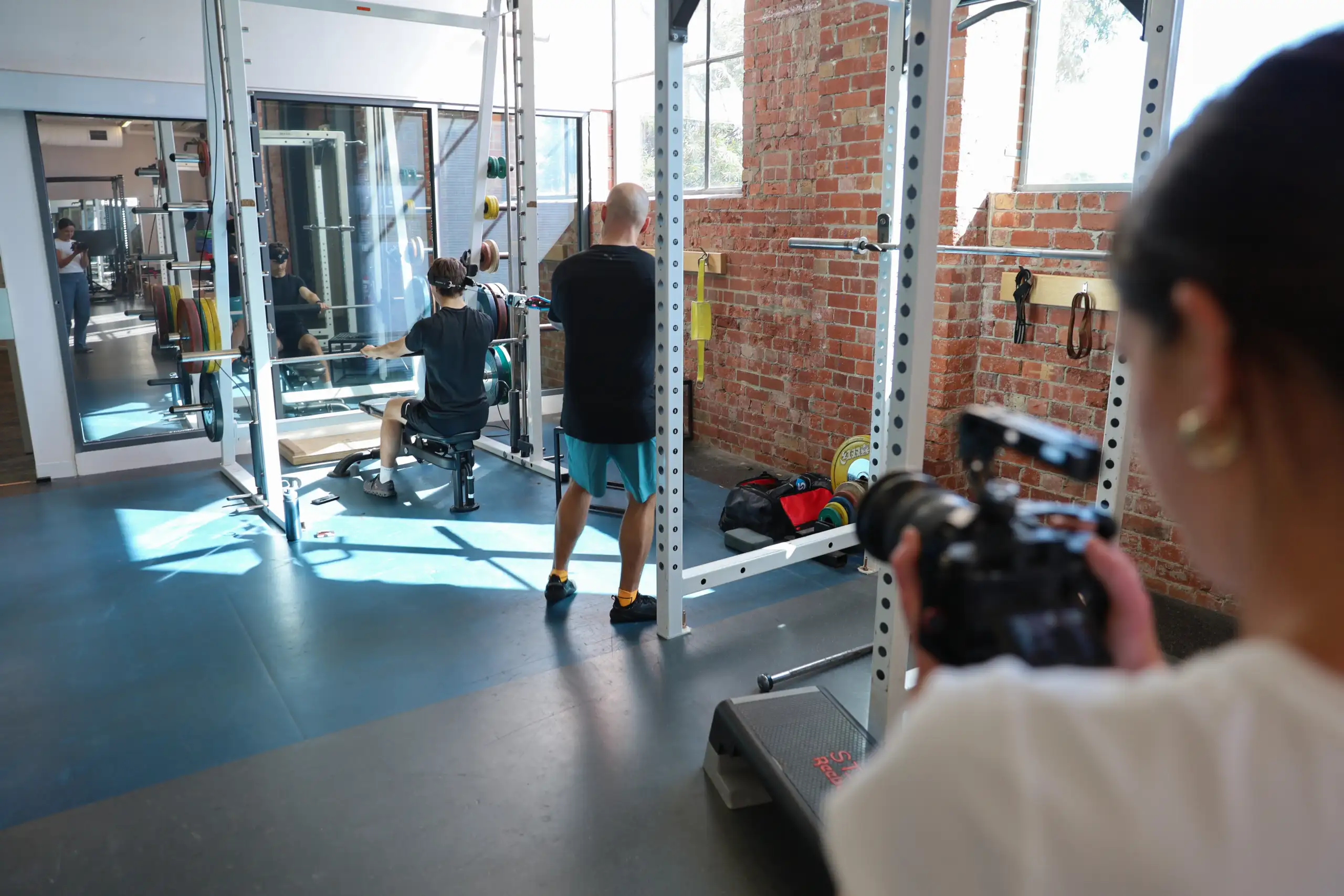 The photo is taken in a gym, in the background is an athlete performing an exercise. In the foreground is the back of a young female in a white t-shirt with a camera in her hands pointing at the athlete.