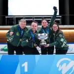 Four Caucasian adults, two male and two female, all wearing Australian team branded jumpers in dark green, stand behind a blue banner with the number 1 on it, holding a large cup trophy. They all smile to camera with their hands in fists or in the air.